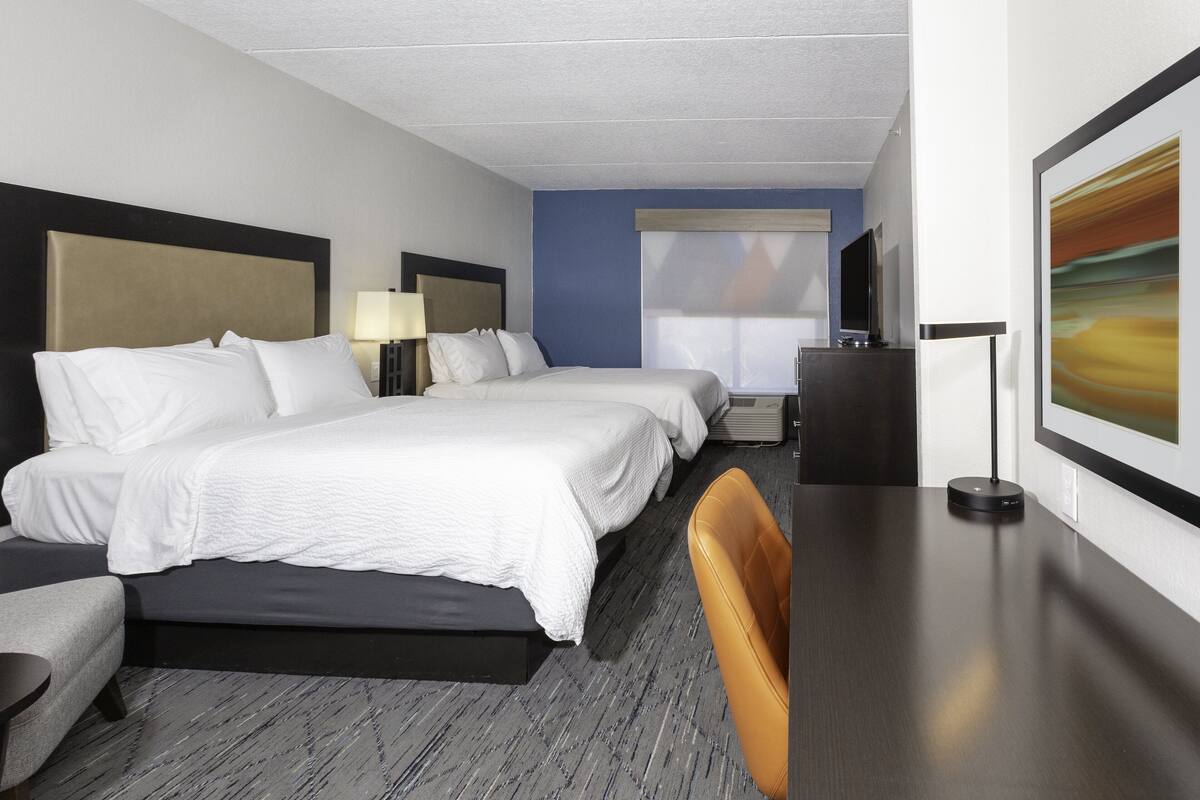 Une chambre d’hôtel avec deux lits, un bureau, une chaise, une télévision et une fenêtre avec des stores.