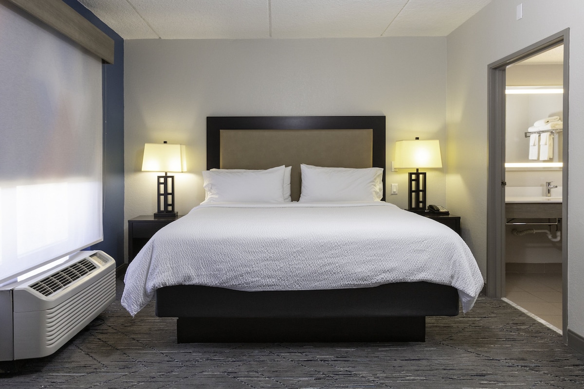 Habitación de hotel con una cama grande, dos mesitas de noche con lámparas, un aire acondicionado instalado en la pared y un baño visible a través de una puerta abierta.