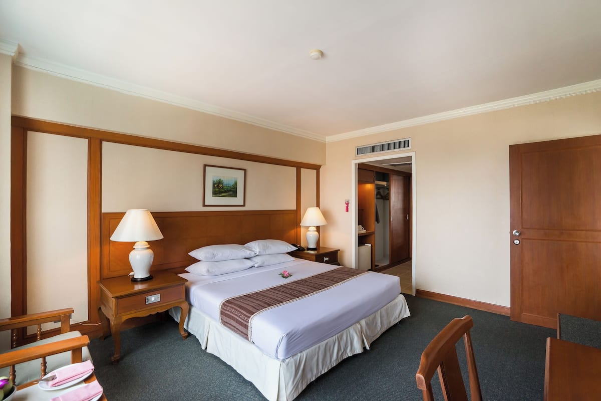 A hotel room with a bed, two bedside tables with lamps, a wooden headboard, a wooden wardrobe, and a small wooden chair.