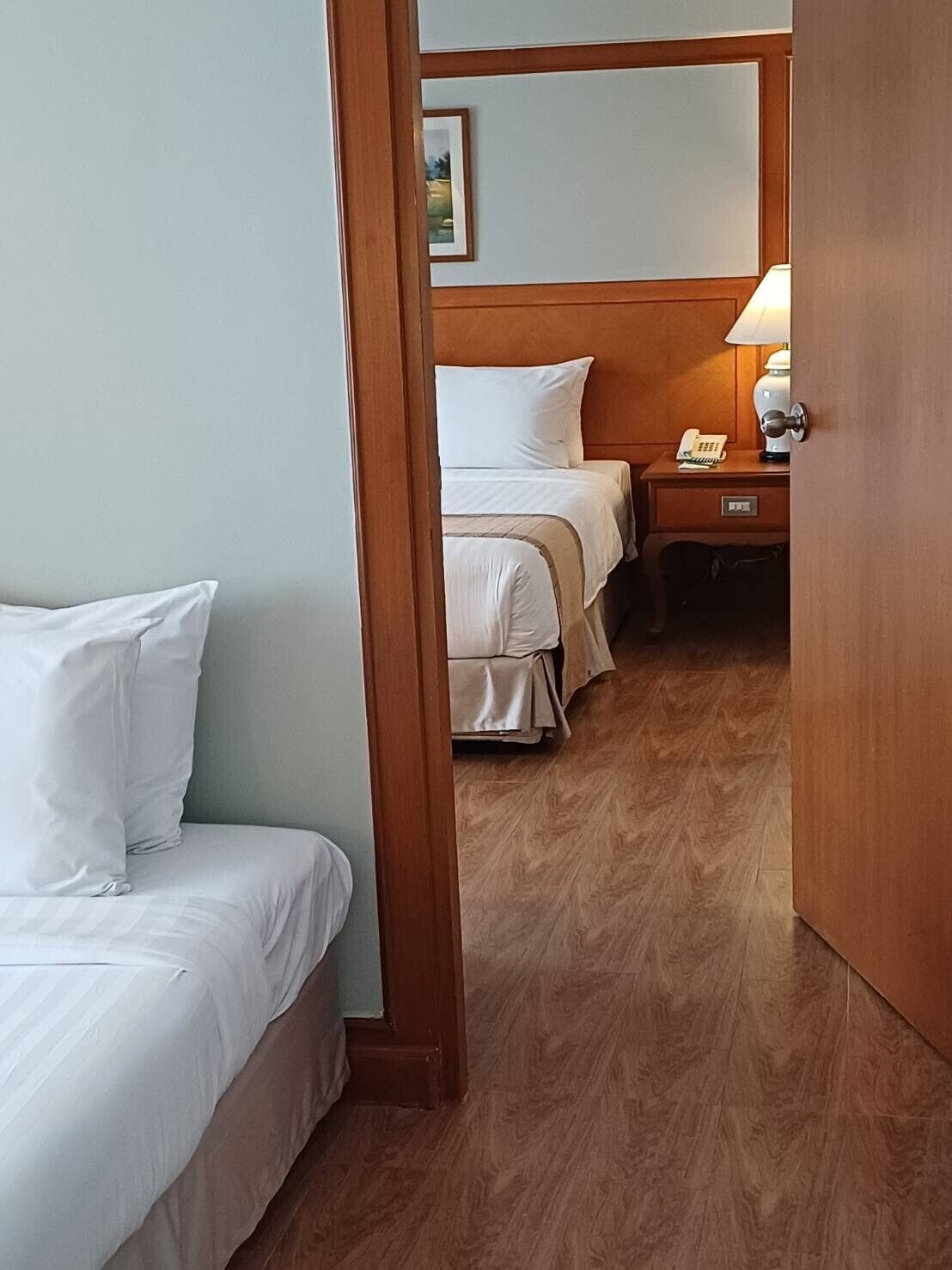 Habitación de hotel con dos camas, cabecero de madera, mesita de noche con lámpara y un cuadro en la pared.