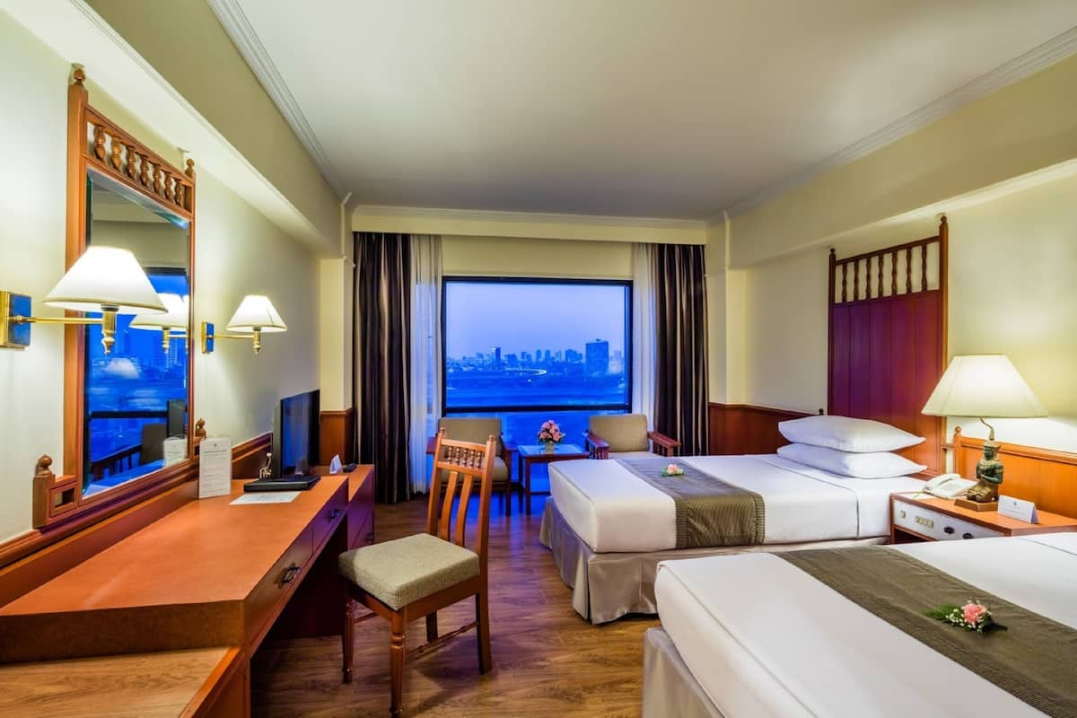 A hotel room with a desk, two beds, a chair, and a large window with a city view.