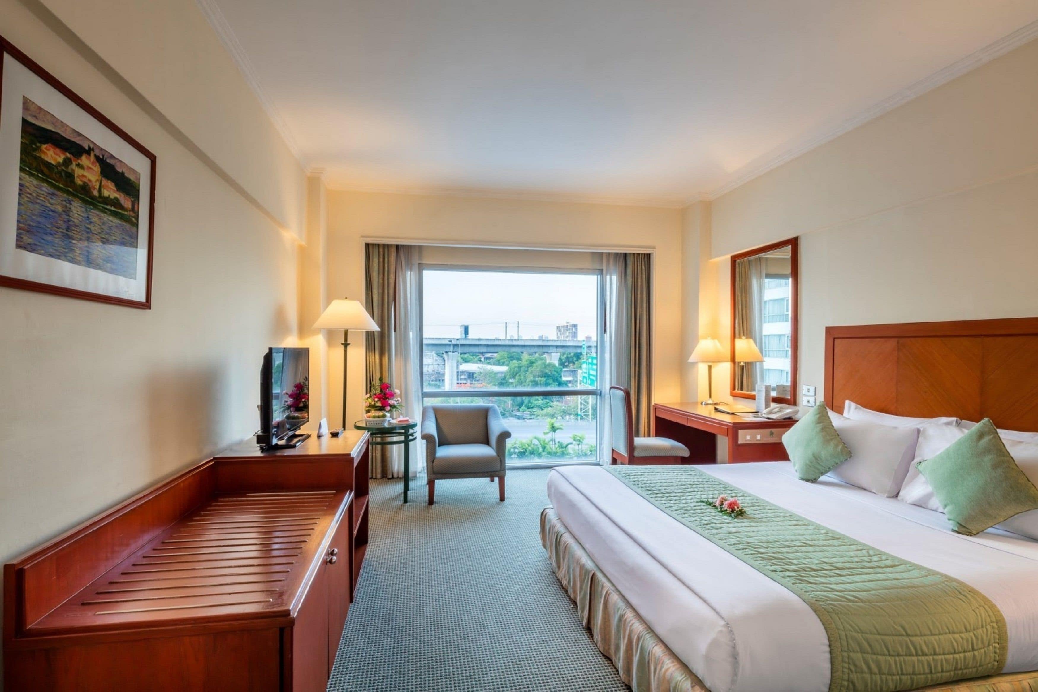 A hotel room with a bed, a chair, a desk, a lamp, a window with a view, and a painting on the wall.