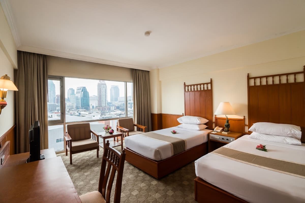A hotel room with two beds, a desk, a chair, and a view of the cityscape.