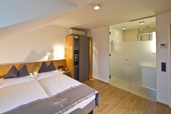 Habitación de hotel con cama, mesita de noche, lámpara, armario y una puerta de vidrio que da a un baño.