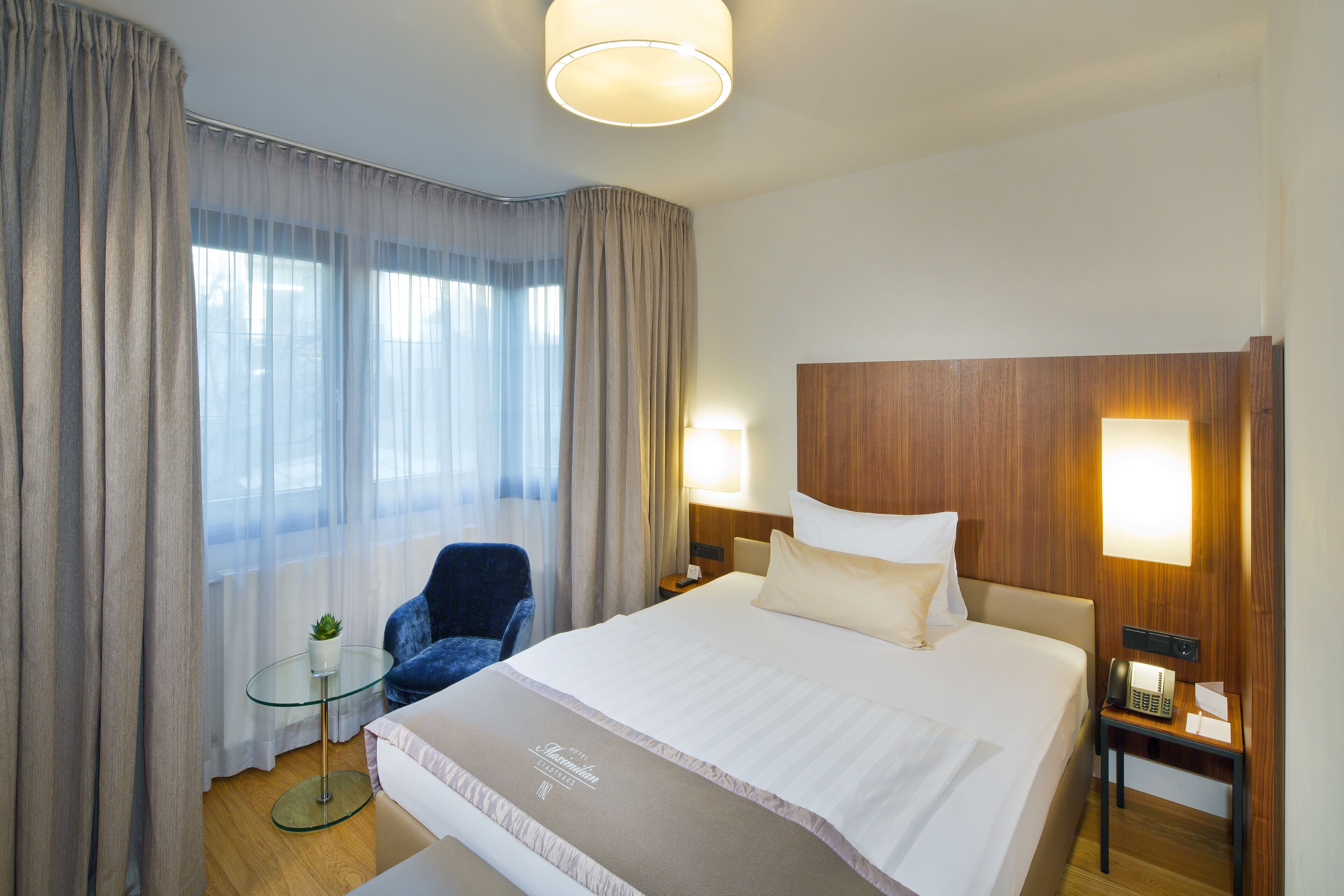 A hotel room with a large bed, a bedside table with a lamp, a chair, and a small table with a plant.