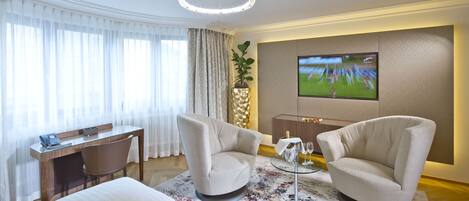 Habitación de hotel con escritorio, dos sillones, televisor de pantalla plana, planta en maceta y alfombra decorativa.