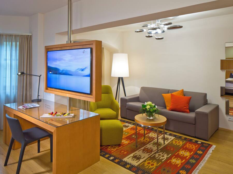 A modern hotel room with a wooden desk, a grey sofa, a green armchair, and a TV mounted on the wall.