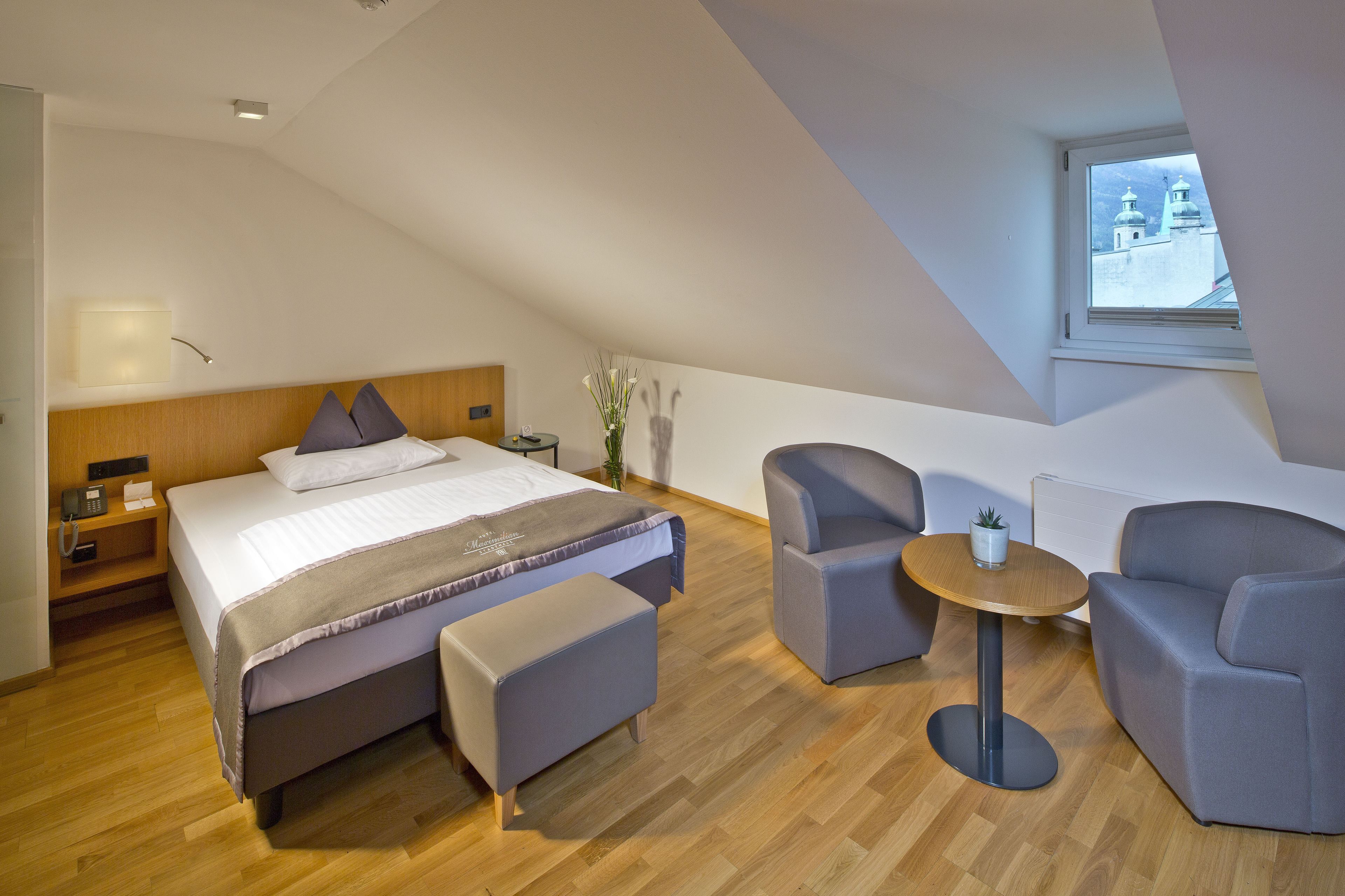 A modern hotel room with a bed, two armchairs, a small round table, and a window with a view of buildings.