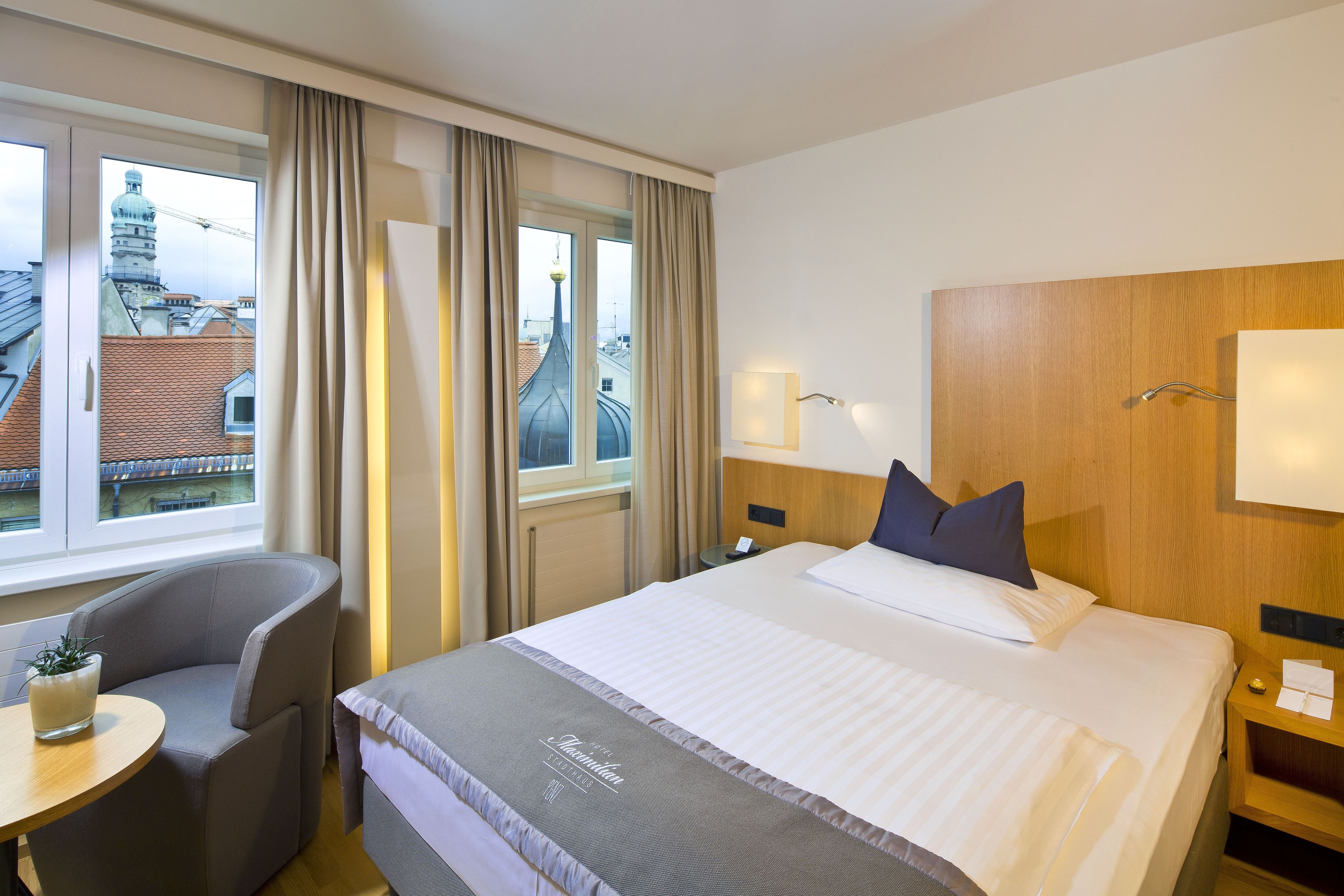 A hotel room with a large bed, a bedside table, a chair, a small table with a plant, and a window with curtains.