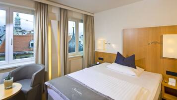 A hotel room with a large bed, a bedside table, a chair, a small table with a plant, and a window with curtains.