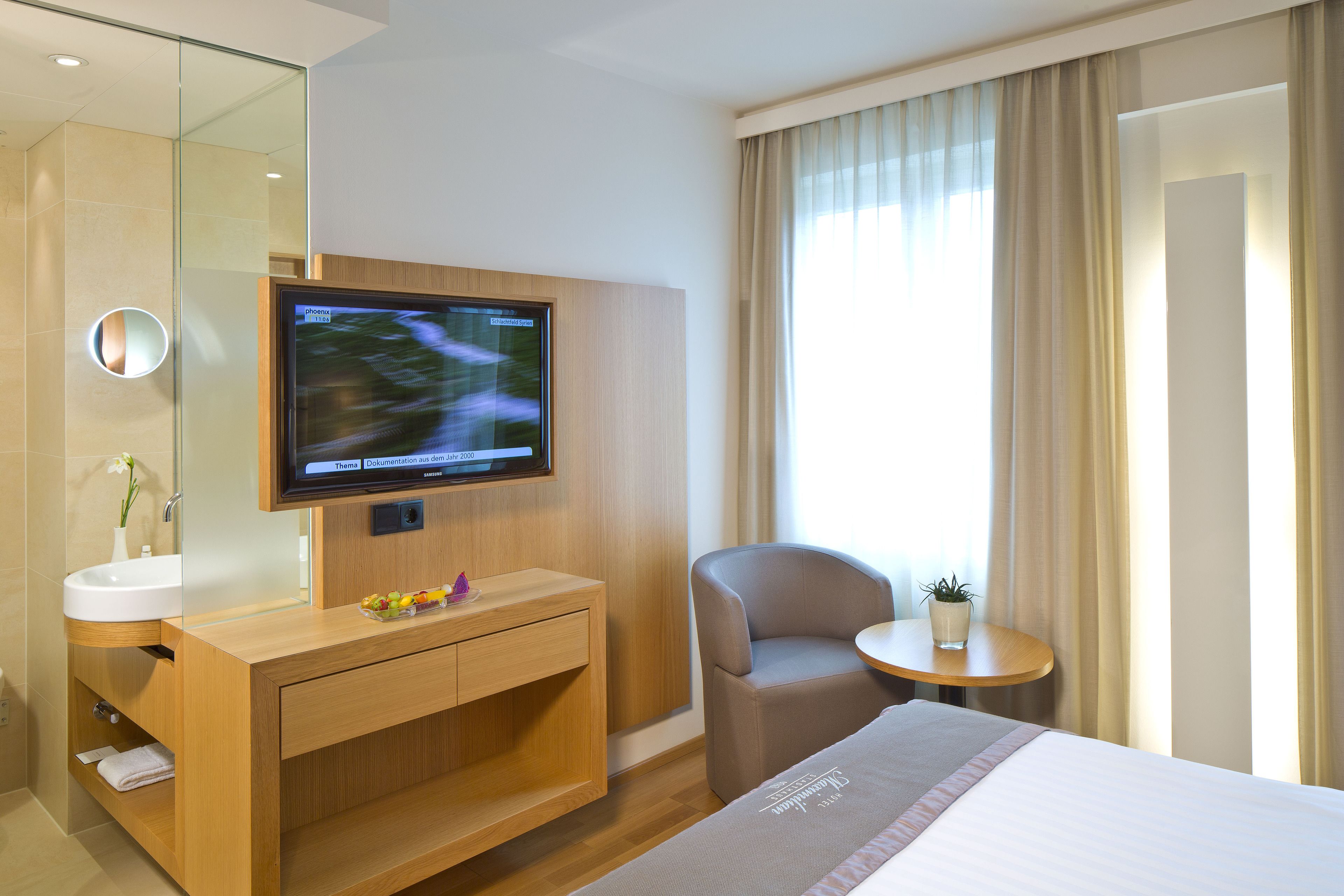 Quarto de hotel com cama, televisão fixada na parede, toucador de madeira com pia, cadeira e pequena mesa com planta.