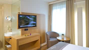 Quarto de hotel com cama, televisão fixada na parede, toucador de madeira com pia, cadeira e pequena mesa com planta.