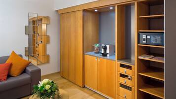 A modern living room with a grey sofa, orange cushions, a wooden coffee table with a floral arrangement, a built-in wardrobe with mirrored panels, and a built-in kitchen area with a microwave and a coffee machine.