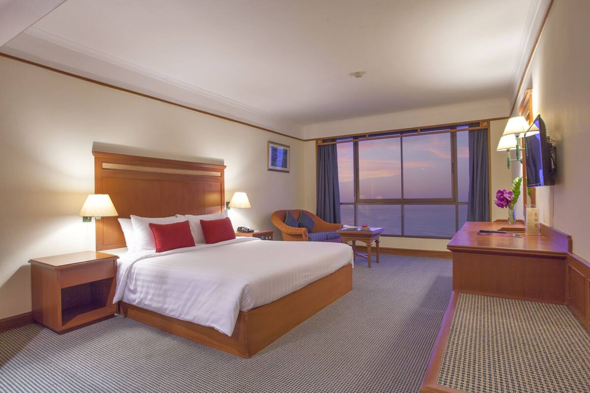 Quarto de hotel com uma cama grande, duas mesas de cabeceira com luminárias, uma cadeira, uma escrivaninha e uma janela ampla com vista para o céu.