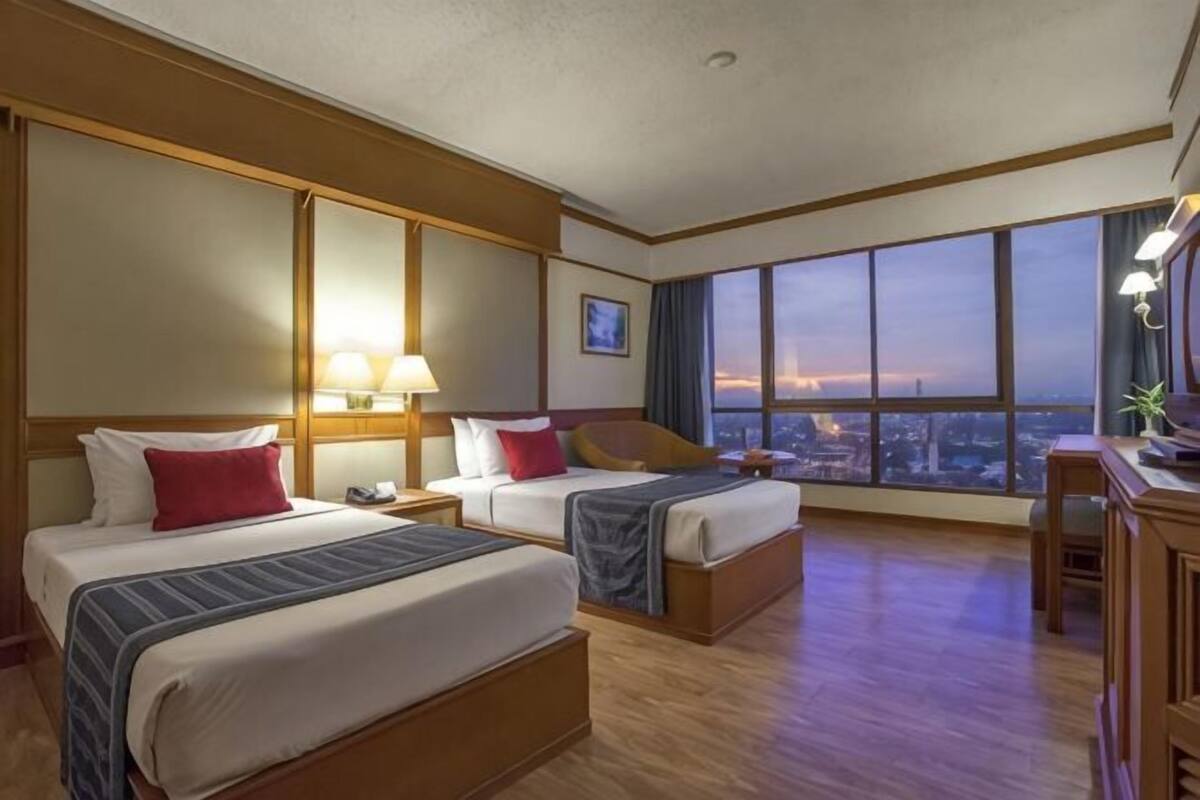Quarto de hotel com duas camas, uma mesa, uma cadeira e uma janela grande com vista para a cidade.