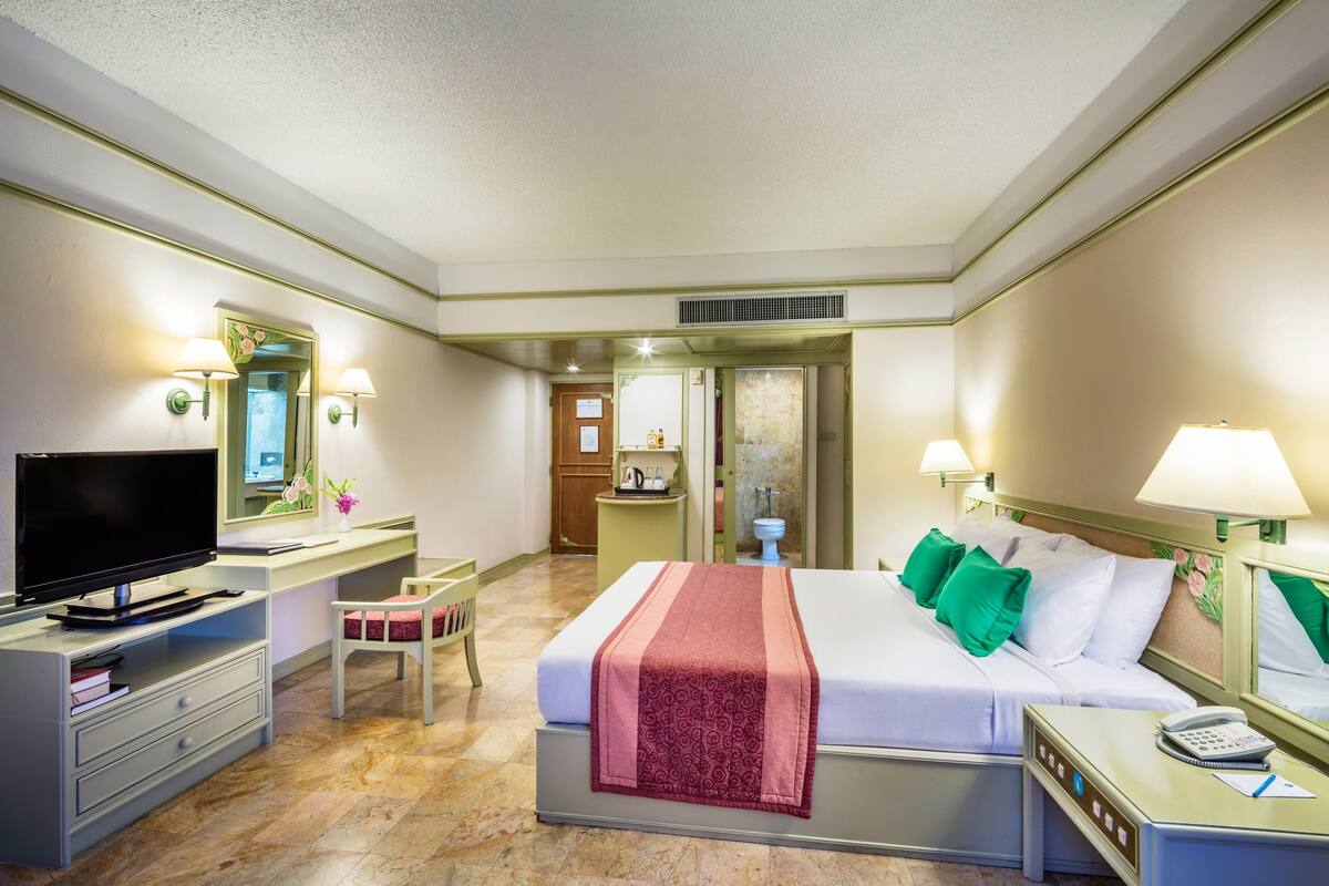 Quarto de hotel com cama, mesa de cabeceira, televisão, escrivaninha, cadeira e banheiro visível por uma porta aberta.