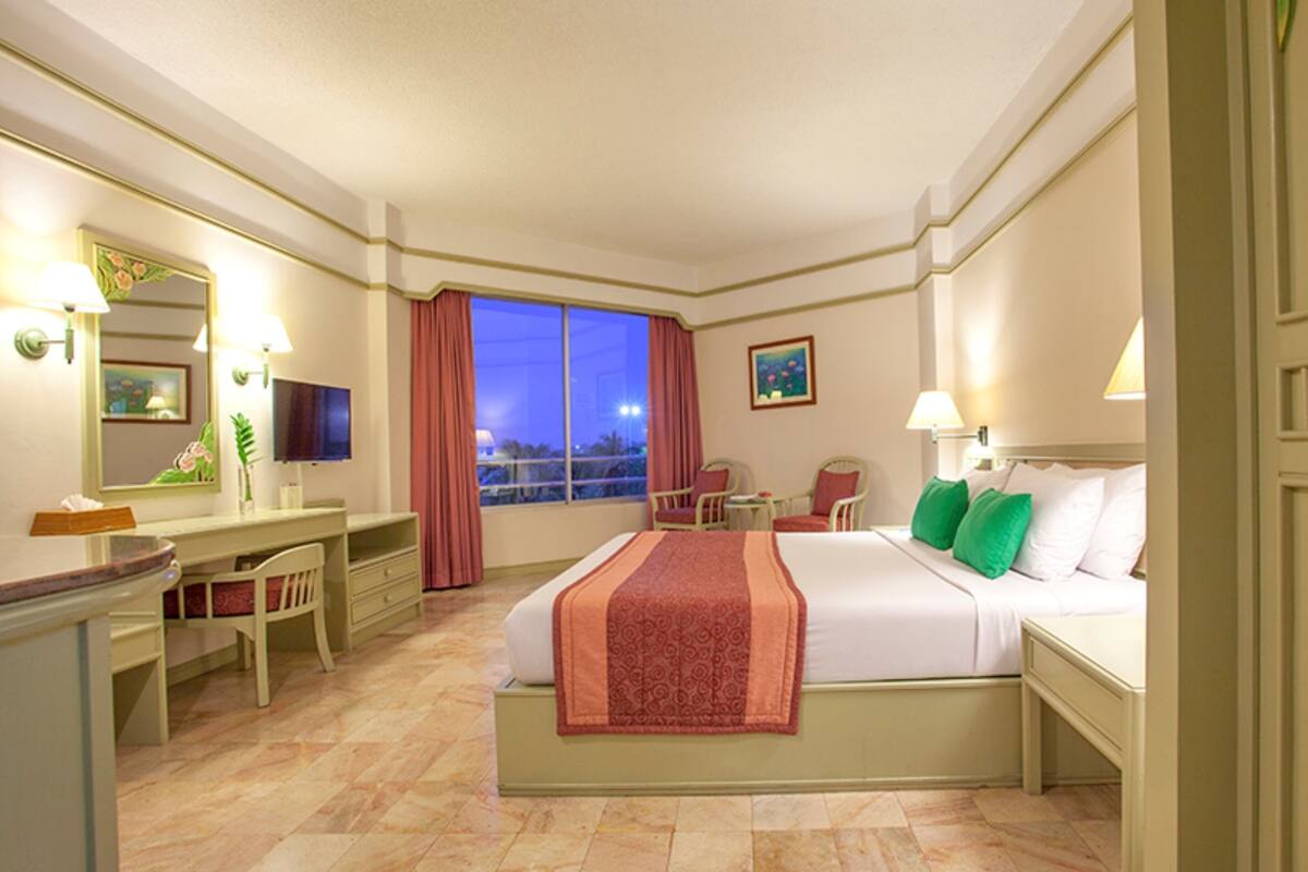 Quarto de hotel com cama, escrivaninha, cadeira e uma janela grande.
