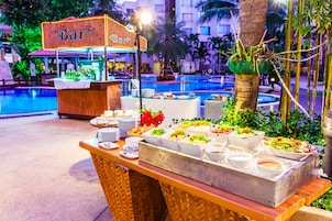 Zona de bufé con un letrero que dice 'Bar', diferentes platos de comida y una piscina al fondo.