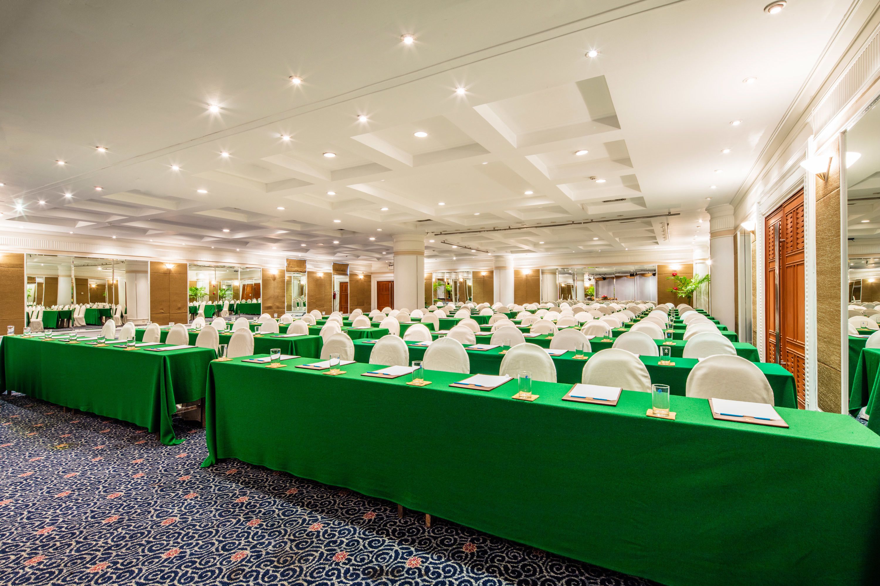 Un salón de conferencias con filas de mesas, manteles verdes, sillas blancas y una alfombra estampada.