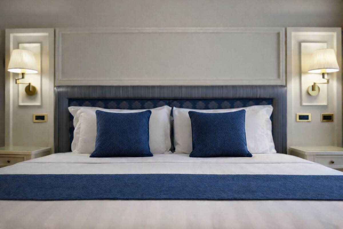 A hotel room with a large bed, two matching blue pillows, a white bedspread, and two wall-mounted lamps on either side of the bed.