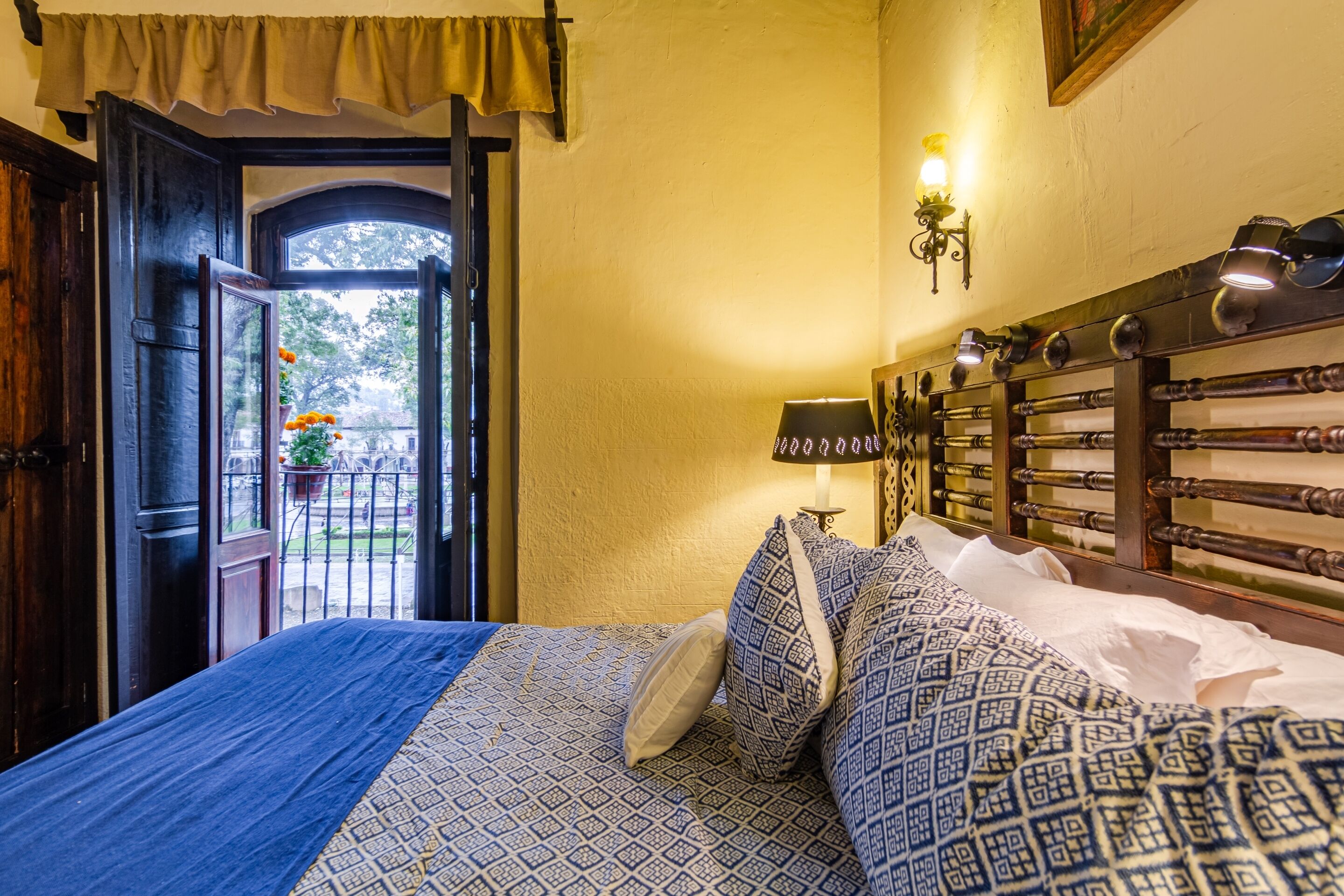 Um quarto com cama, janela com cortinas, abajur e luminária na parede.