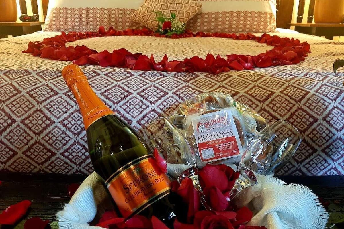 Un lit avec des coussins à motifs, une bouteille de champagne, une bouteille de Morellanas et un bouquet de roses rouges.