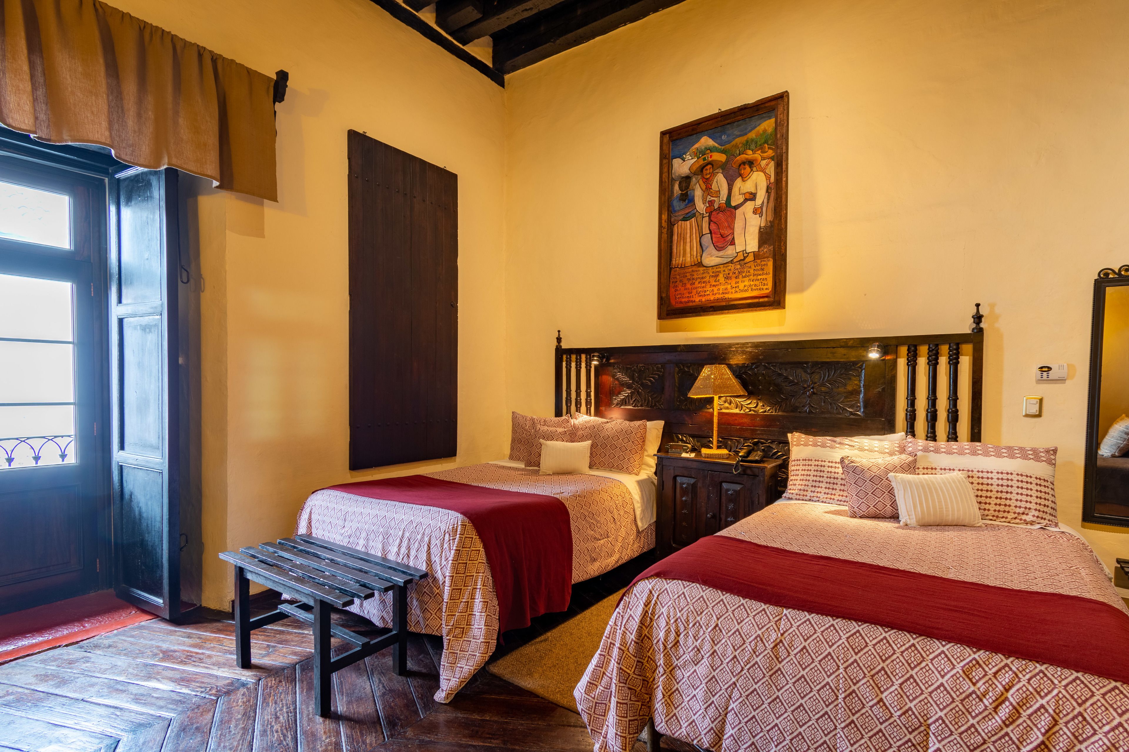 Habitación de hotel con dos camas, un banco, una puerta de madera y un cuadro en la pared.