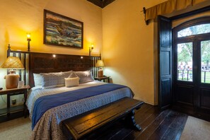 Un dormitorio ordenado con una cama grande, dos lámparas de noche, un banco, un cuadro en la pared y vistas a través de una puerta abierta.