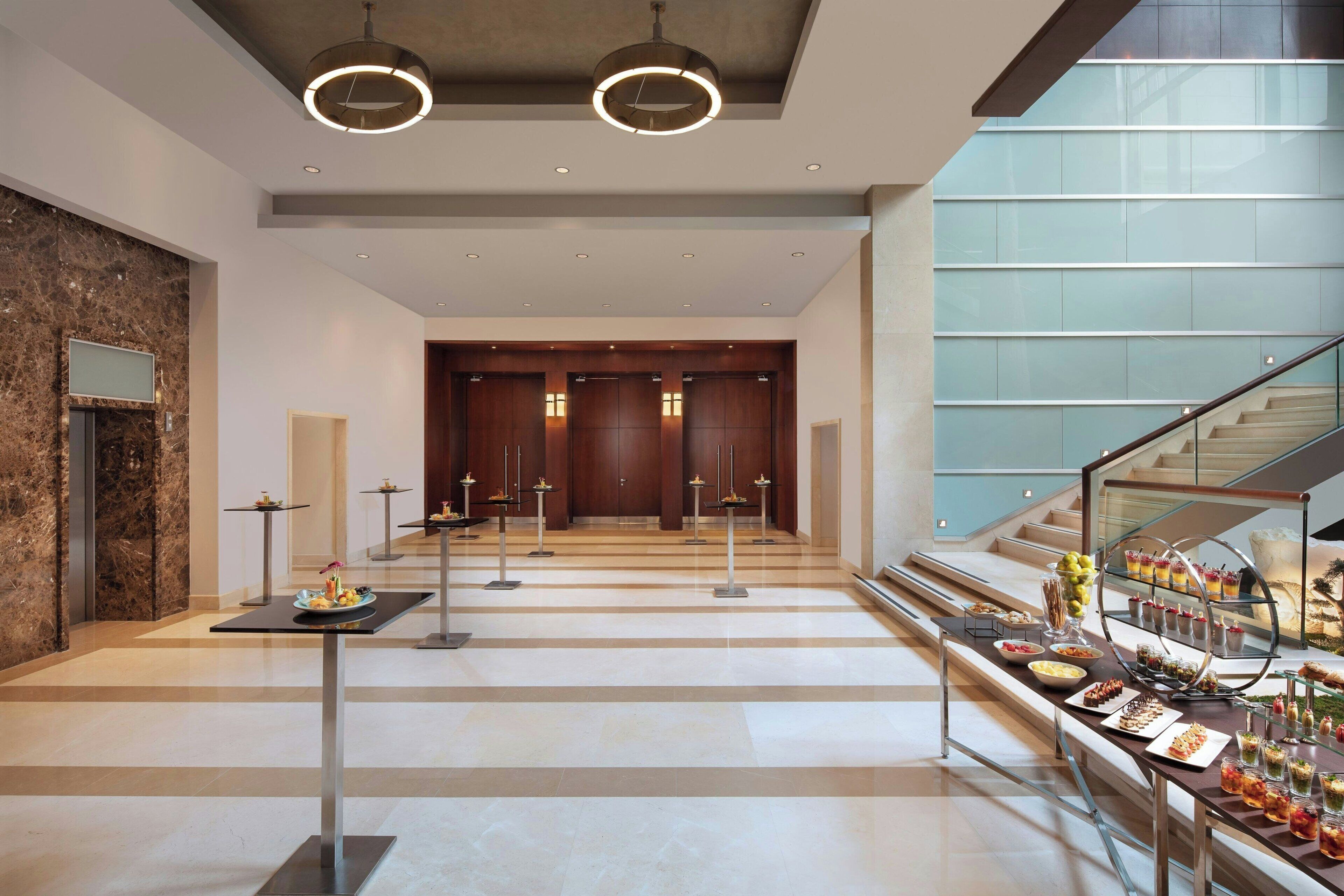 A modern hotel lobby with a marble floor, a reception desk, a staircase, and a buffet area.
