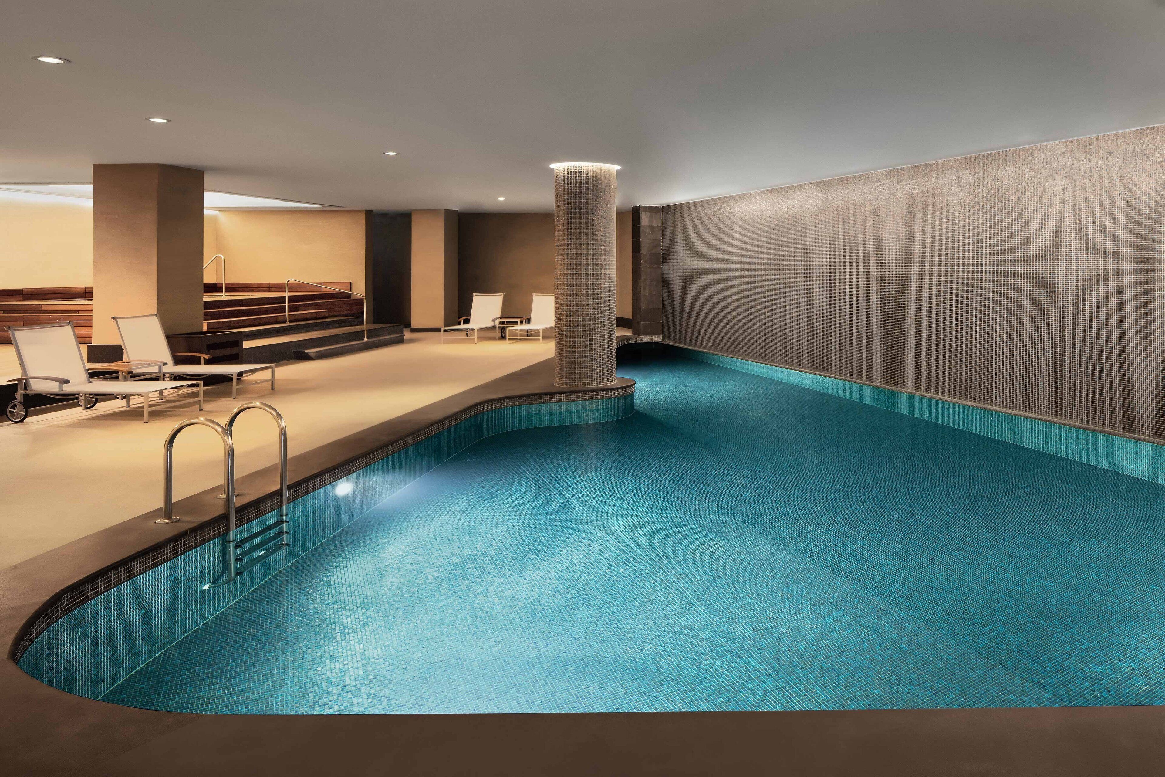 A modern indoor swimming pool with a clear blue water, a central column, and lounge chairs along the poolside.