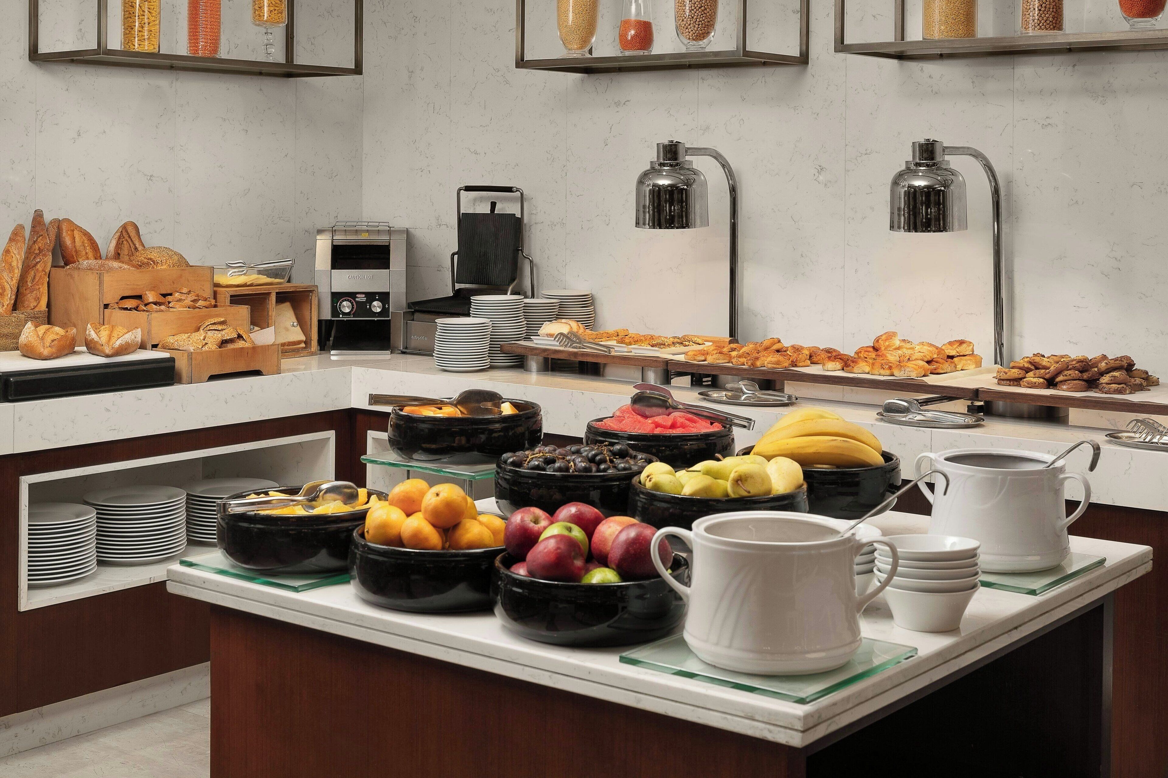 A buffet with various food items, including fruits, vegetables, and pastries. There are also several bowls and plates, as well as a coffee machine and a lamp.