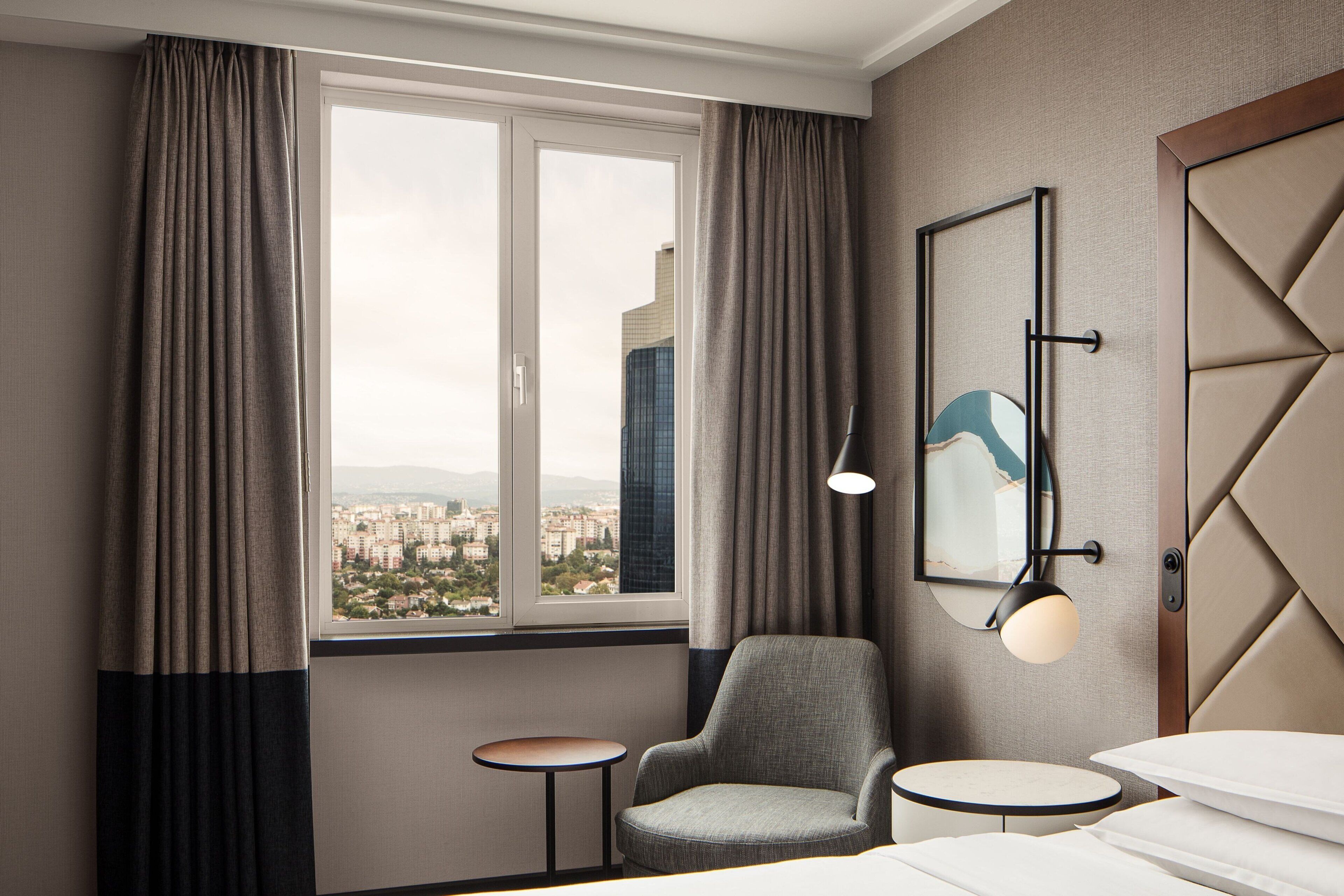 A hotel room with a large window, a chair, a small table, and a bed with pillows.