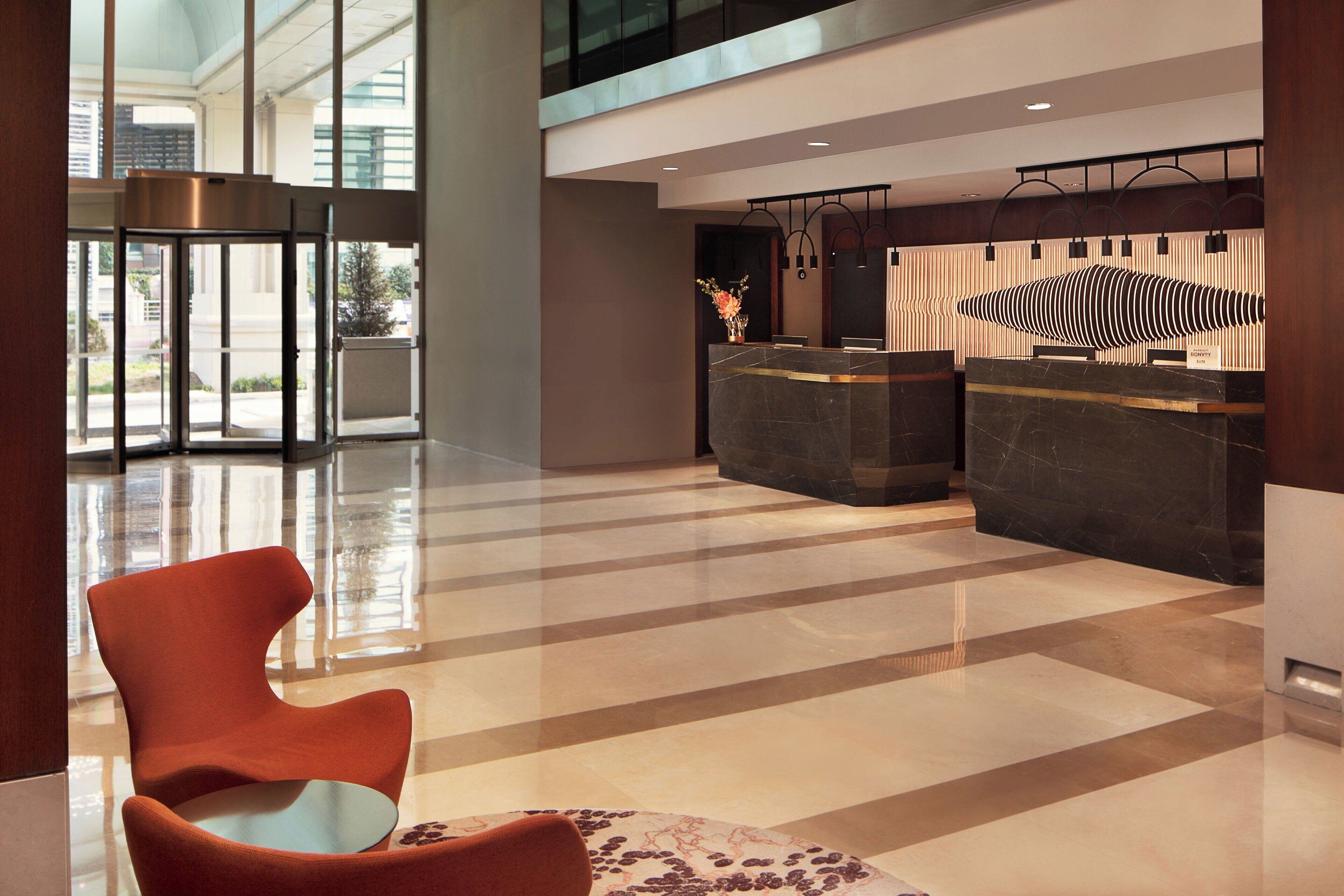 A modern lobby with a reception desk, a seating area with a round table, and a large glass door leading outside.