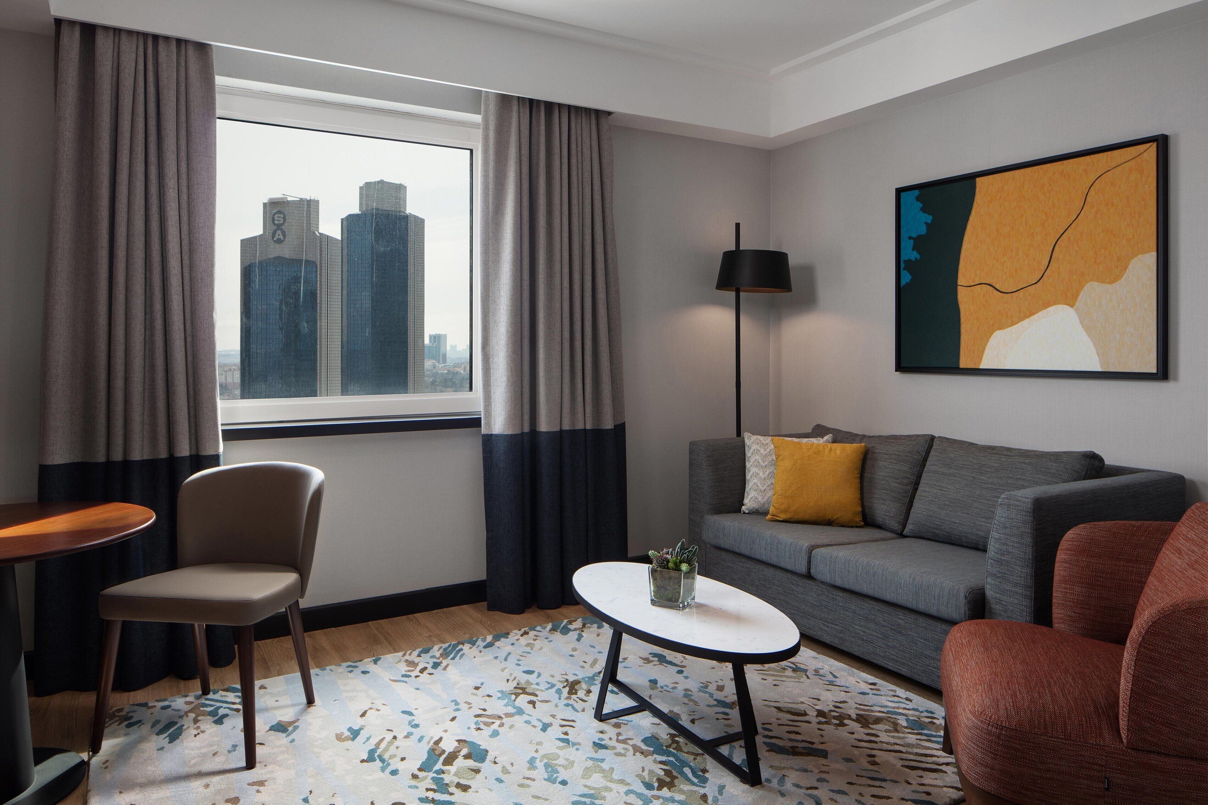 A modern hotel room with a city view, a grey sofa, a red armchair, a small table, and a floor lamp.