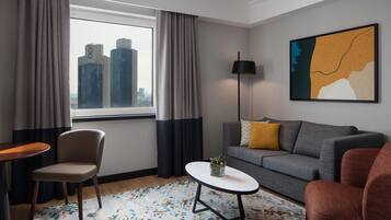 Una habitación de hotel moderna con vistas a la ciudad, un sofá gris, un sillón rojo, una mesita y una lámpara de pie.