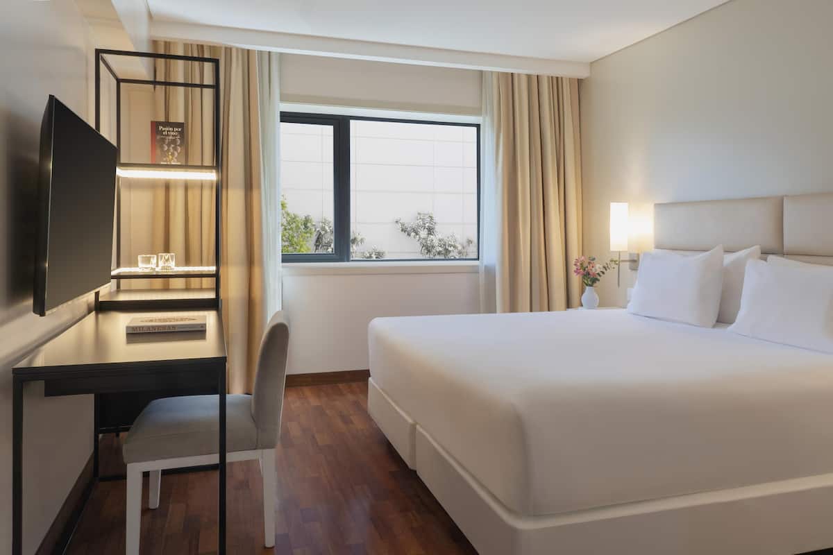 A hotel room with a bed, a desk with a chair, a TV, a window with curtains, and a nightstand with a lamp and flowers.