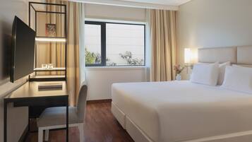 Habitación de hotel con cama, escritorio y silla, televisión, ventana con cortinas y mesita de noche con lámpara y flores.