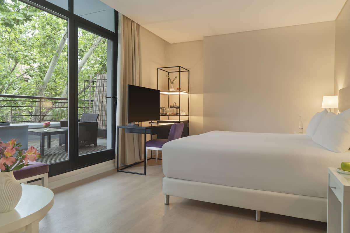 A hotel room with a large bed, a desk with a computer, a chair, a small table with a vase, and a balcony with a table and chairs.
