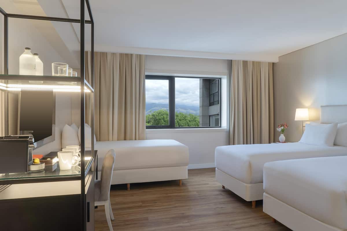 A hotel room with two beds, a desk with a laptop, a chair, a large window with curtains, and a view of greenery outside.