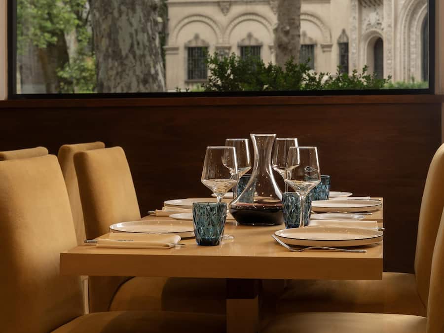 Mesa de comedor dispuesta con platos, vasos y un florero, vista a través de una ventana que da a un árbol y un edificio con balcones.