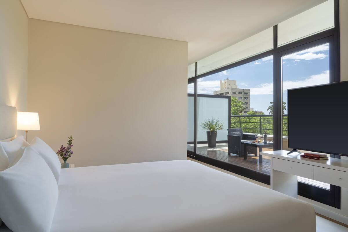 A modern hotel room with a large bed, a TV, and a balcony with a view of buildings and greenery.