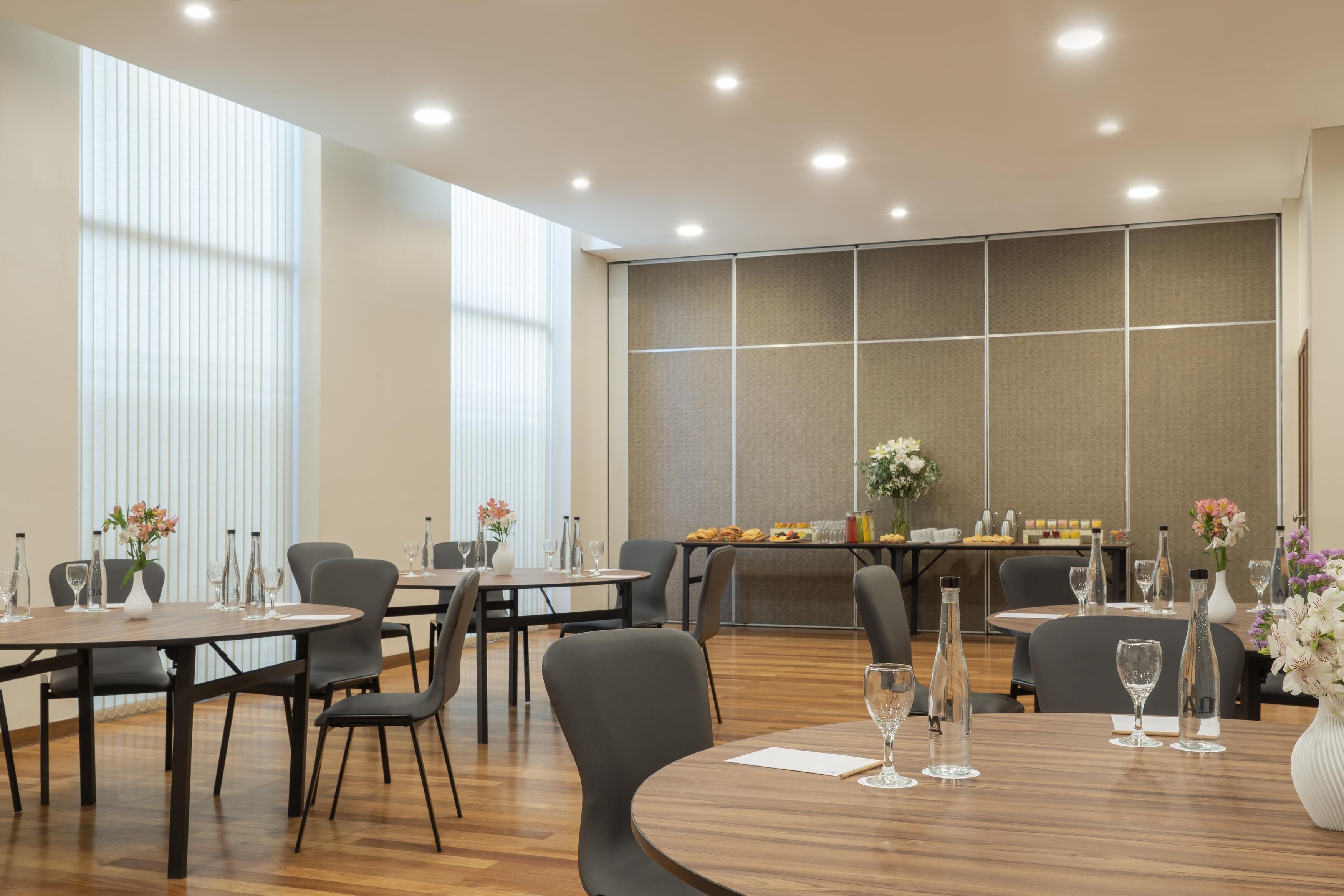 A modern dining area with round tables, grey chairs, and a long table with a buffet.