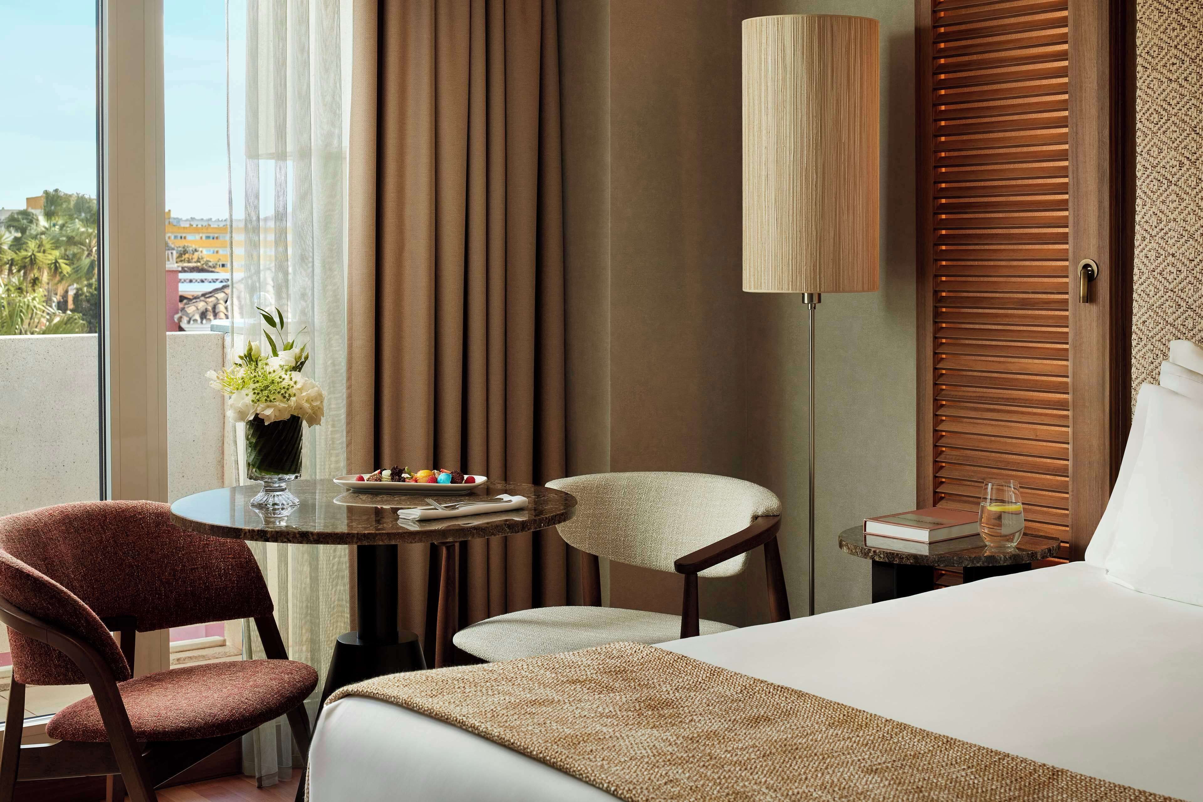 Habitación de hotel con una cama, dos sillas, una mesita con un florero y una lámpara, y vistas al exterior.
