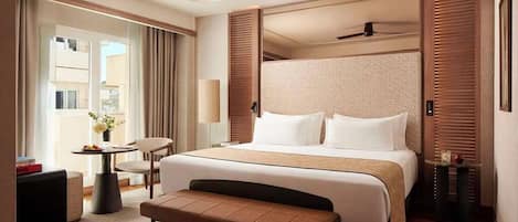 Habitación de hotel moderna con una cama grande, un ventilador de techo, una pequeña mesa con una lámpara, un banco y vistas al exterior.