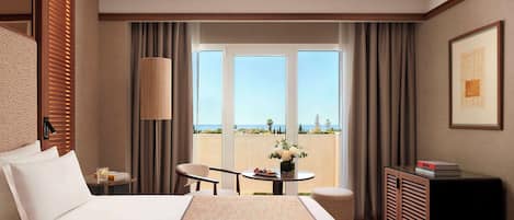 Habitación de hotel con una cama grande, un escritorio, una silla, una mesita con un florero y vistas al exterior a través de amplias ventanas.