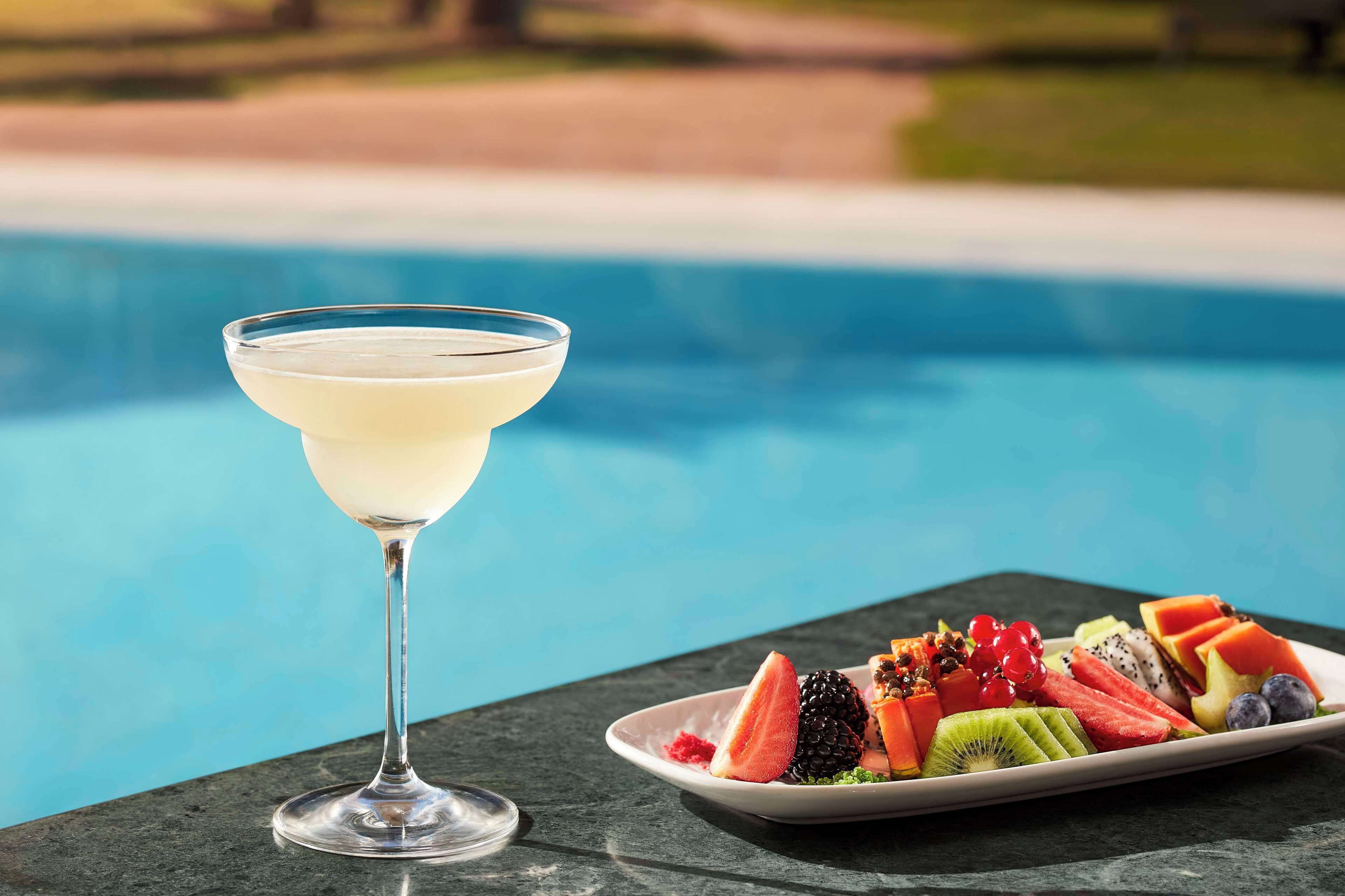 Un verre à martini posé à côté d’une assiette de fruits variés, près d’une piscine.