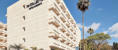 A modern hotel building with a prominent logo, multiple balconies, and a well-maintained landscaped area with palm trees.