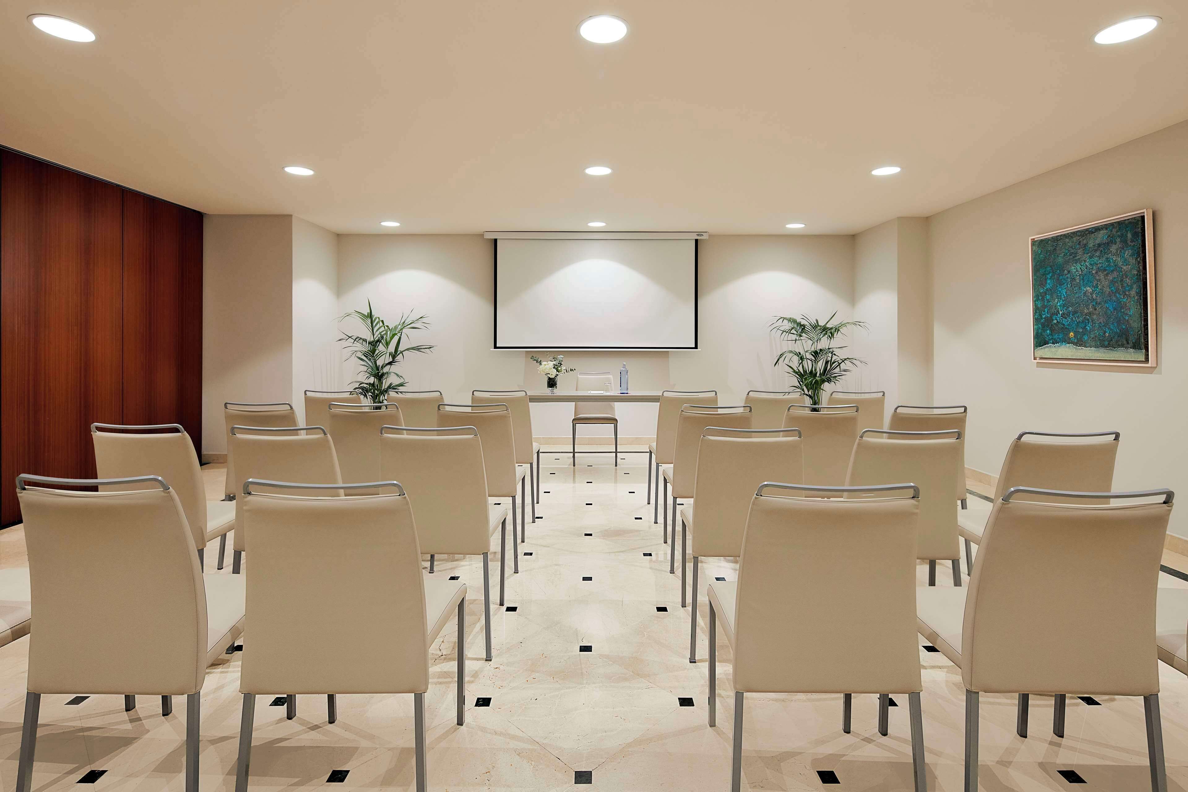 A conference room with rows of chairs facing a central screen, wooden paneling on one side, and a painting on the wall.