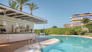Zona de piscina con bar, palmeras y un edificio al fondo.