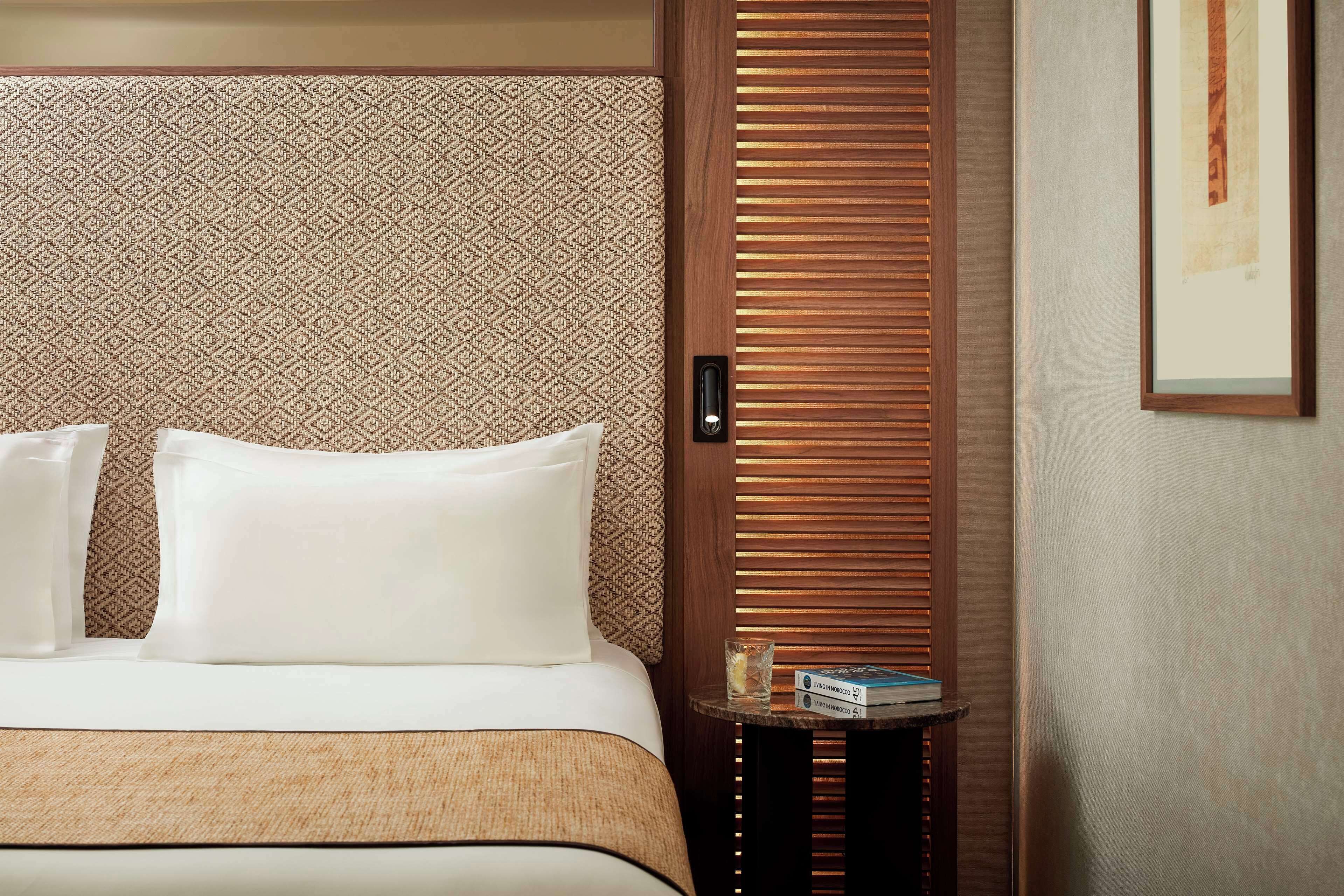 A hotel room with a bed, pillows, a small table with a book, and a wooden door with a patterned curtain.
